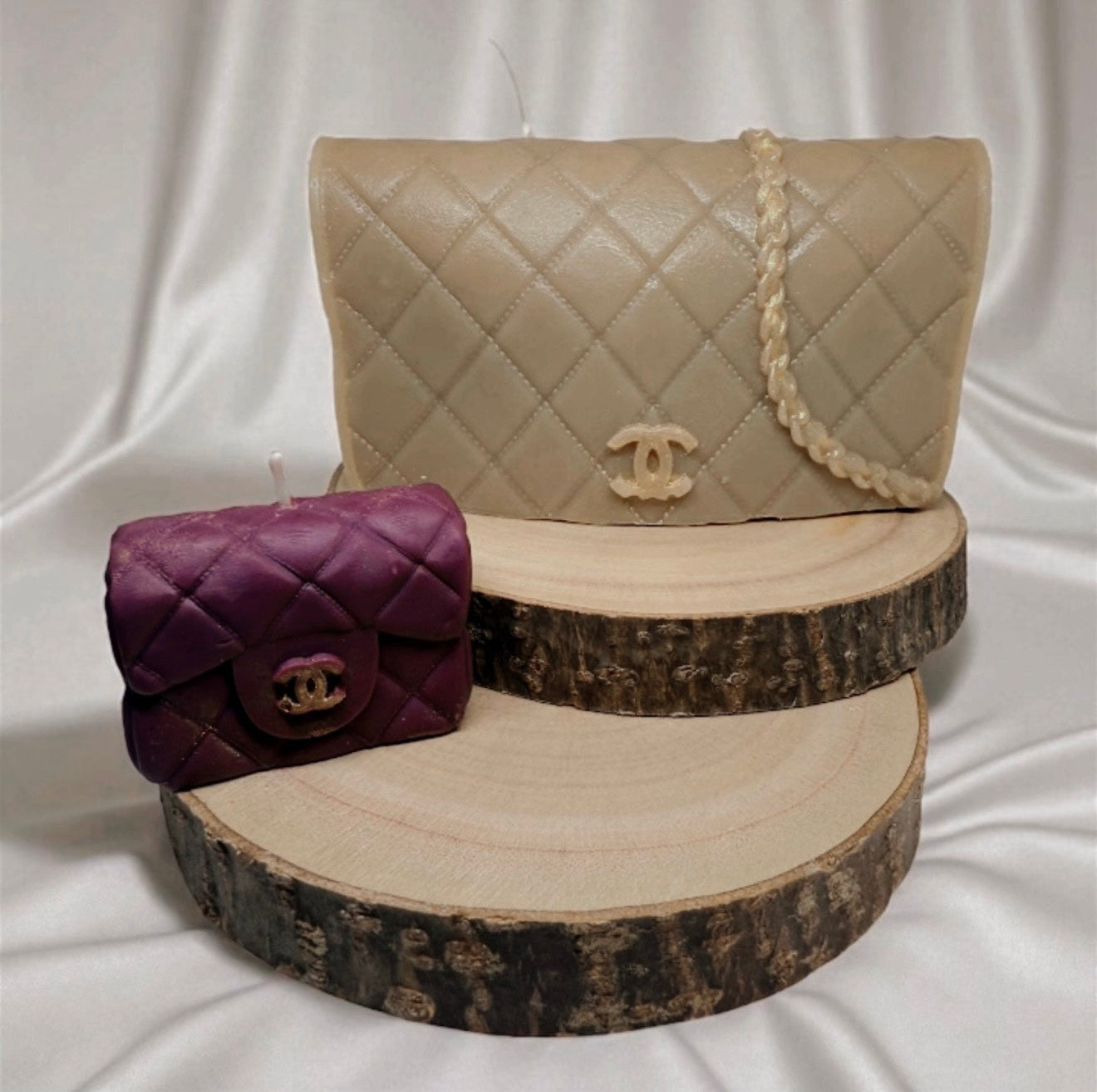 Two luxe couture purse candles inspired by designer handbags, one beige and one purple, displayed on wooden slices against a satin background